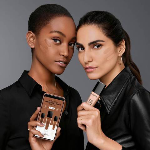 Two women with foundation swatches on their cheeks and holding up Lancome’s e-shade finder color match tool and foundation