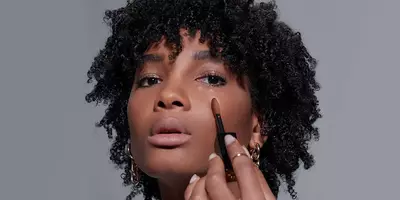Woman with dark skin tone showing how to apply all over concealer under the eyes.